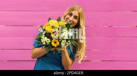 Überraschte Frau in blauem Bademantel präsentiert einen Haufen Frühlingsblumen. Rosafarbener Holzhintergrund Stockfoto