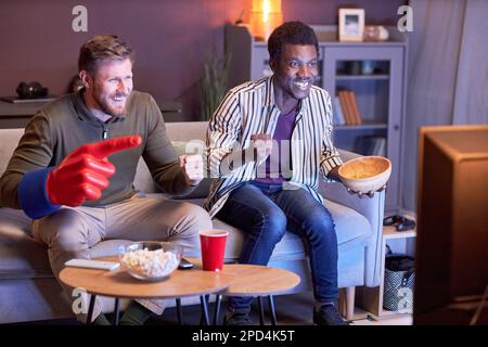 Porträt zweier emotionaler Sportfans, die in blauem Licht Fernsehen und Snacks essen Stockfoto