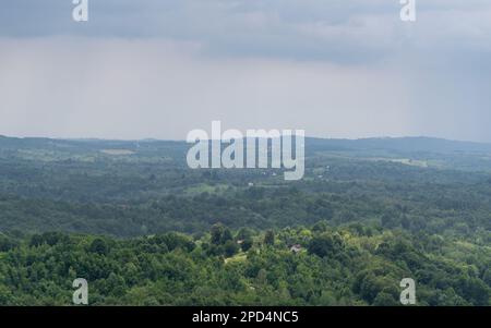 Ländliche Landschaft im Sommer mit verschiedenen Wetterbedingungen, Sonnenfleck auf einem Bauernhof umgeben von üppigen Wäldern und verstreuten Dörfern mit düsterer Atmosphäre Stockfoto