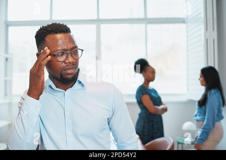Der Schwarze sieht verärgert aus, während Kollegen hinter seinem Rücken reden, Bürodrama Stockfoto