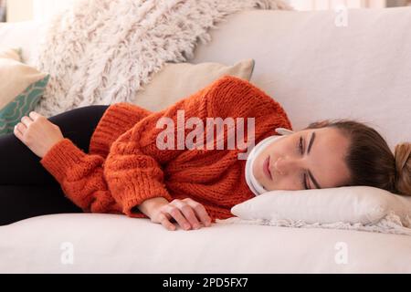 Müde junge Frau in orangefarbener warmer Kleidung mit orthopädischem Kragen, die sich nach einer Zervixverletzung im Wohnzimmer zu Hause auf der Couch ruht Stockfoto