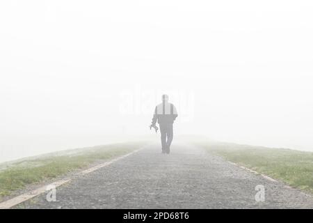 Rückansicht eines einsamen Mannes, der einen langen Weg entlang geht und in den frühen Morgennebel/Nebel verschwindet, der einen Hund in der Hand hält, ohne einen Hund! Stockfoto