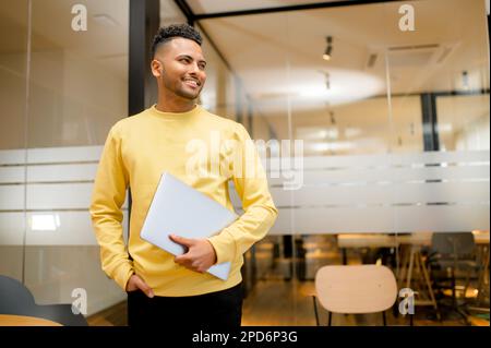Hochqualifizierter, ehrgeiziger Büroangestellter mit Laptop, der in einem modernen Gemeinschaftsraum steht, lächelnder arabischer Student mit Computer, der in der Bibliothek steht, mit Glastrennwänden im Hintergrund Stockfoto