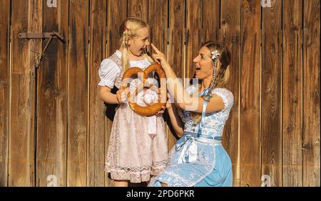 Glückliche Mutter und Tochter, die eine große Brezel in der traditionellen bayerischen Tracht in Bayern auf Holzhintergrund halten. Oktoberfest-Konzeptbild Stockfoto