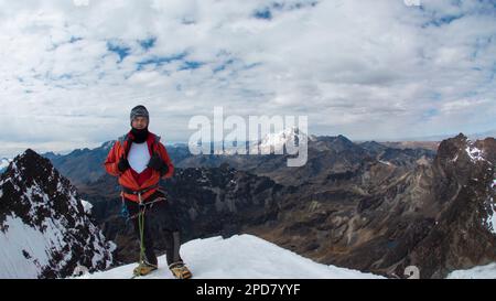 Junger, männlicher Kletterer, der an einem bewölkten Tag auf dem Gipfel des verschneiten Berges mit Helm, Stirnlampe, rotem Mantel und Seil steht Stockfoto