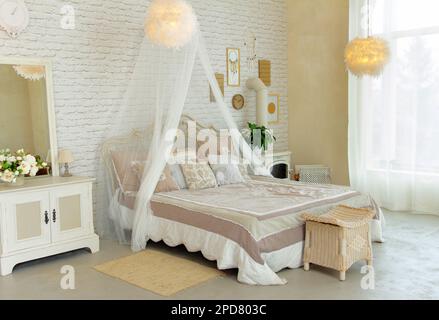 Komfortables, großes, einfaches Schlafzimmer mit hellen Möbeln, Himmelbett, Federlampen, kleinem Kamin auf Holz, gemütlichem, modernem Platz zum Ausruhen und Stockfoto