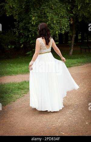 Braunes Mädchen steht in einem wunderschönen, weißen, langen Kleid mit tiefem Schnitt auf dem Rücken, in einem grünen Park Stockfoto