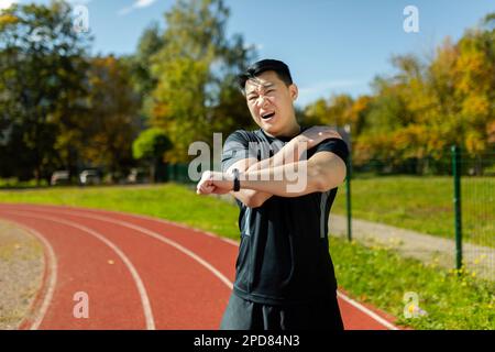 Der asiatische Sportler dehnt sich die Schulter, der Mann befindet sich nach dem Laufen im Stadion und bei sportlichen Übungen dehnt er seine Armgelenke, hat Schulterschmerzen, Muskelkrämpfe. Stockfoto