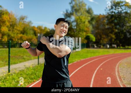 Der asiatische Sportler dehnt sich die Schulter, der Mann befindet sich nach dem Laufen im Stadion und bei sportlichen Übungen dehnt er seine Armgelenke, hat Schulterschmerzen, Muskelkrämpfe. Stockfoto