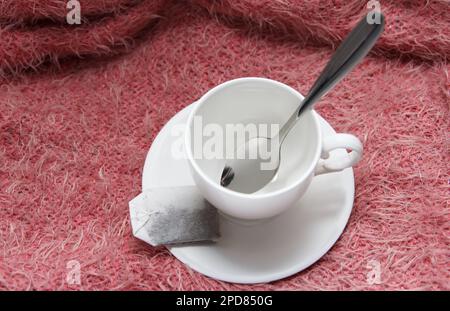 Ein weißer, leerer Becher mit Teelöffel und eine infundierte Tüte daneben in einer Untertasse liegen auf pinkfarbenem Hintergrund. Warten Sie, bis kochendes Wasser eingegossen wird Stockfoto