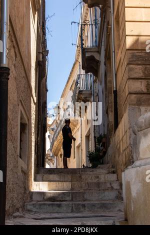 In Scicli, Italien, auf der 06-08-22, steht ein Mann auf der Treppe in einer typischen Straße des historischen Zentrums Stockfoto