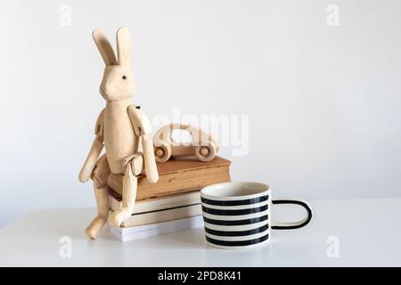 Stillleben mit einem Holzhasen auf Scharnieren im Innenraum. Ein weiß-schwarz gestreifter Becher, ein Stapel Bücher und ein hölzernes Kinderspielzeugauto. Speicherplatz kopieren Stockfoto