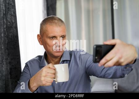 Ein Mann mittleren Alters mit Tee macht ein Selfie-Foto mit einem Handy in der Wohnung. Stockfoto