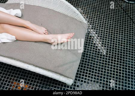 Nahaufnahme der Unterschenkel einer nicht wiedererkennbaren jungen Frau im weißen Bademantel entspannt auf einem Liegestuhl am Pool nach einer Schönheitsbehandlung im Stockfoto