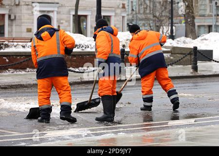 Drei Arbeiter säubern Schnee mit Schaufeln, Schneeräumen in der Stadt. Frühlingswetter, Straßenreinigung Stockfoto