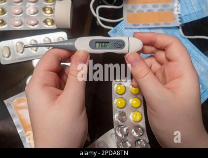 Elektronisches Thermometer in den Händen vor dem Hintergrund eines medikamentösen Tischs (Nahaufnahme). Konzept von Medizin, Apotheke, Gesundheitswesen und häuslicher Behandlung Stockfoto