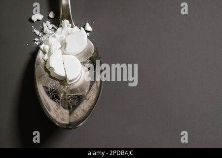 Pharmazeutisches Arzneimittelpulver auf einem silbernen Metalllöffel. Zerschmettertes Mittel, Tabletten, oben auf einem grauen Tisch im Hintergrund. Stockfoto