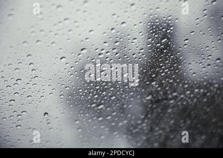 Regentropfen auf einem Fenster mit einer sanft verschwommenen Stadtlandschaft dahinter, die eine stimmungsvolle, atmosphärische Szene erzeugen. Ideal für Wetter, melancholische, gemütliche Momente in Innenräumen, Stockfoto