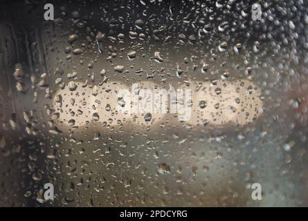 Regentropfen auf einem Fenster mit einer sanft verschwommenen Stadtlandschaft dahinter, die eine stimmungsvolle, atmosphärische Szene erzeugen. Ideal für Wetter, melancholische, gemütliche Momente in Innenräumen, Stockfoto