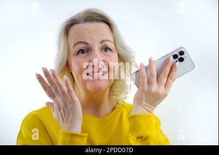Überraschung peinliche Schande Frau in orangefarbenem gelben Pullover steht in Händen mit Telefon schüchtern schließt Augen rötete Gesichtsklammern Zähne berühren Kopf, um überrascht zu sein peinlich welche Art von Nachricht Stockfoto