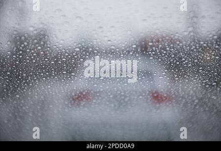 Regentropfen auf einem Fenster mit einer sanft verschwommenen Stadtlandschaft dahinter, die eine stimmungsvolle, atmosphärische Szene erzeugen. Ideal für Wetter, melancholische, gemütliche Momente in Innenräumen, Stockfoto