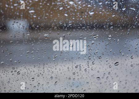 Regentropfen auf einem Fenster mit einer sanft verschwommenen Stadtlandschaft dahinter, die eine stimmungsvolle, atmosphärische Szene erzeugen. Ideal für Wetter, melancholische, gemütliche Momente in Innenräumen, Stockfoto
