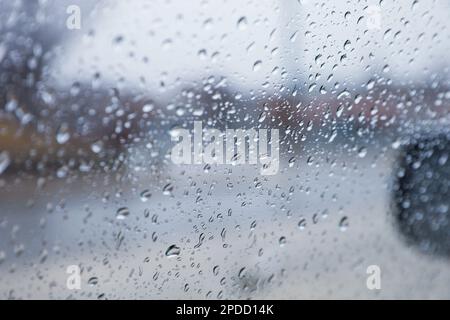 Regentropfen auf einem Fenster mit einer sanft verschwommenen Stadtlandschaft dahinter, die eine stimmungsvolle, atmosphärische Szene erzeugen. Ideal für Wetter, melancholische, gemütliche Momente in Innenräumen, Stockfoto