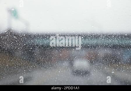 Regentropfen auf einem Fenster mit einer sanft verschwommenen Stadtlandschaft dahinter, die eine stimmungsvolle, atmosphärische Szene erzeugen. Ideal für Wetter, melancholische, gemütliche Momente in Innenräumen, Stockfoto