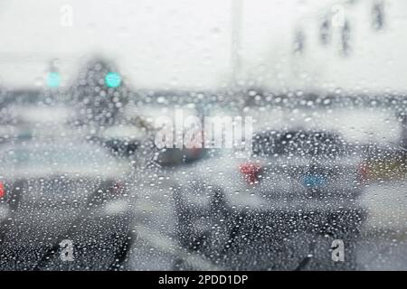 Regentropfen auf einem Fenster mit einer sanft verschwommenen Stadtlandschaft dahinter, die eine stimmungsvolle, atmosphärische Szene erzeugen. Ideal für Wetter, melancholische, gemütliche Momente in Innenräumen, Stockfoto