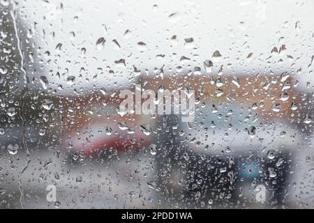 Regentropfen auf einem Fenster mit einer sanft verschwommenen Stadtlandschaft dahinter, die eine stimmungsvolle, atmosphärische Szene erzeugen. Ideal für Wetter, melancholische, gemütliche Momente in Innenräumen, Stockfoto