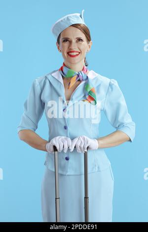 Lächelnde, elegante Flugbegleiterin mit blauem Hintergrund in blauer Uniform und Reisetasche, die nach oben in den Kopierbereich schaut. Stockfoto