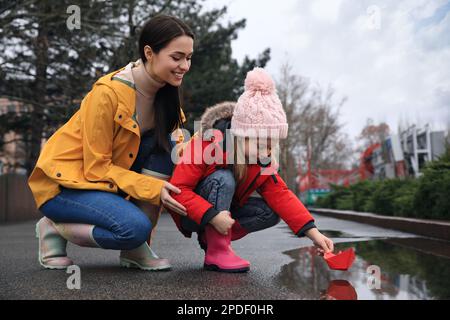 Das kleine Mädchen und ihre Mutter spielen mit einem Papierboot in der Nähe der Pfütze im Freien Stockfoto