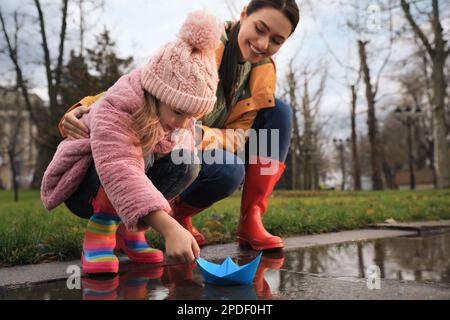 Das kleine Mädchen und ihre Mutter spielen mit einem Papierboot in der Nähe der Pfütze im Park Stockfoto