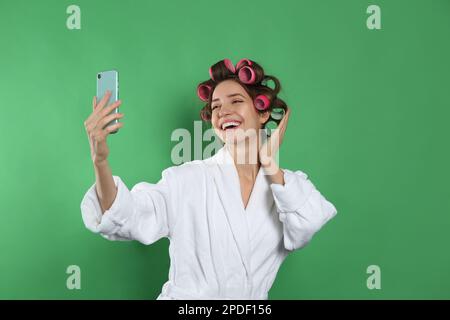 Glückliche junge Frau im Bademantel mit Lockenwicklern, die Selfie auf grünem Hintergrund macht Stockfoto