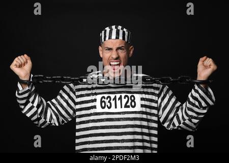 Emotionaler Gefangener in gestreifter Uniform, der versucht, die Kette auf schwarzem Hintergrund zu brechen Stockfoto