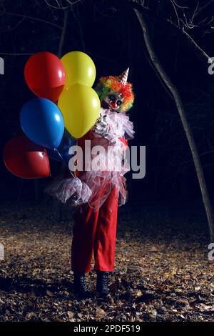 Furchteinflößender Clown mit Luftballons draußen in der Nacht. Halloween-Party-Kostüm Stockfoto