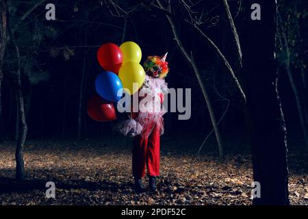 Furchteinflößender Clown mit Luftballons draußen in der Nacht. Halloween-Party-Kostüm Stockfoto