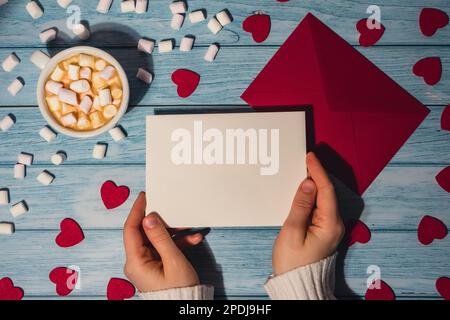 Weibliche Hände mit leerer valentinskarte, roter Umschlag mit weißer Tasse Kaffee und Marshmallows auf blauem Holzhintergrund. Romantische kleine Herzen Valentinstag. Leerer Papierkartenkopierplatz für Ihren Text. Feiertagmorgen. Ansicht von oben, flach liegend, minimalistisches Branding-Konzept Stockfoto