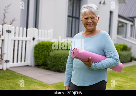Porträt einer lächelnden, birassischen Seniorin mit kurzen grauen Haaren, die Yoga-Matte im Hof hält, Kopierraum Stockfoto