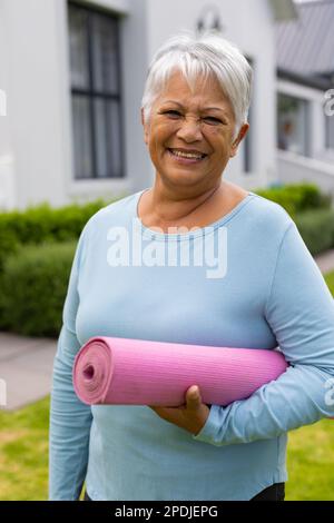 Lächelnde, birassische Seniorin mit kurzen grauen Haaren, die eine Trainingsmatte gegen das Haus im Hof hält Stockfoto