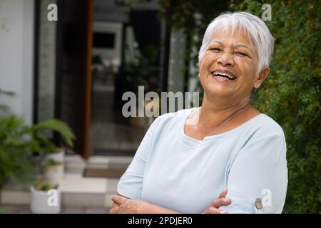 Fröhliche, birassische ältere Frau mit kurzen grauen Haaren, die die Arme kreuzt und vor dem Haus steht Stockfoto