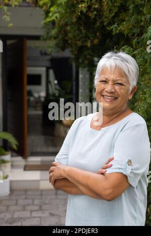 Fröhliche, birassische Seniorin mit kurzen grauen Haaren, die die Arme kreuzen und vor dem Haus neben dem Efeu stehen Stockfoto
