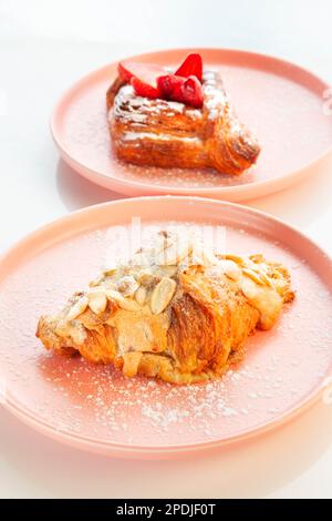 Mandelcroissant auf einem rosafarbenen Teller mit Erdbeer-plunderstückchen im Hintergrund. Stockfoto