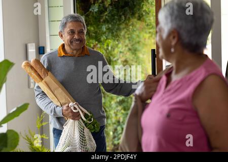 Lächelnder, birassistischer Senior-Mann, der Brot und Lebensmittel in einer Netztasche trug und seine Frau vor der Tür anstarrte Stockfoto
