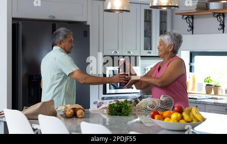 Birassische Seniorin, die ihrem Mann eine Saftflasche gibt, während sie Lebensmittel auf der Küchentheke auspackt Stockfoto