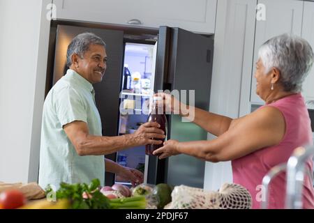 Birassische Seniorin, die einem lächelnden Ehemann eine Saftflasche gibt, steht am Kühlschrank in der Küche Stockfoto