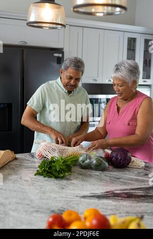 Birassischer älterer Ehemann, der seiner Frau beim Auspacken von Lebensmitteln aus Netzbeuteln auf dem Küchentisch hilft Stockfoto