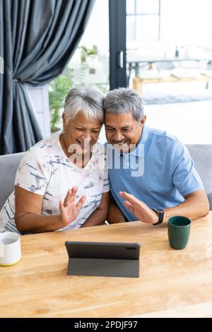 Fröhliches, birassistisches Seniorenpaar, das sich zu Hause mit der Hand winkt, während es sich über Videoanrufe auf einem digitalen Tablet-PC zu Hause unterhält Stockfoto