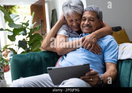 Lächelnde, birassische Seniorin, die ihren Ehemann von hinten umarmt und ein digitales Tablet auf dem Sofa verwendet Stockfoto