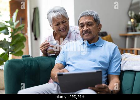Lächelnde, birassische Seniorin mit Kaffeetasse, die sich von ihrem Ehemann zu Hause mit einem Tablet auf dem Sofa lehnt Stockfoto